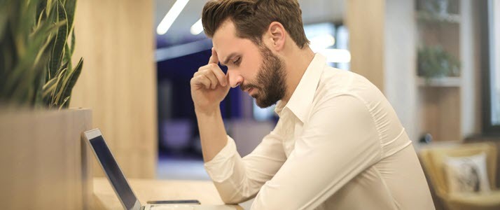 Employee Burnout Signs A man sits with his hand on his temple looking at a laptop in frustration