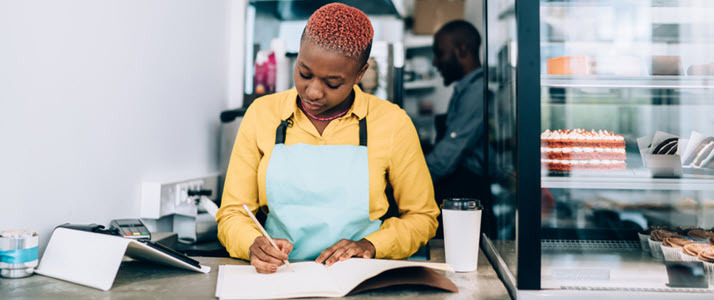 Black woman at bakery counter writing notes in notebook Employee Handbooks10 Must-Have Policies for 2022