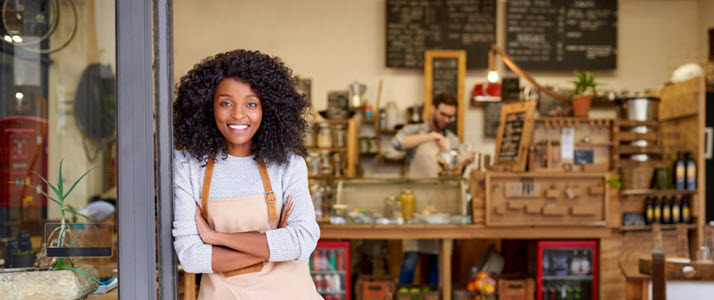 A smiling young Black business owner leaning with her arms crossed on the door of a cafe