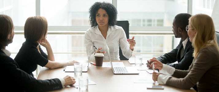 A woman professional leads her colleagues in discussion as they all sit around a conference table