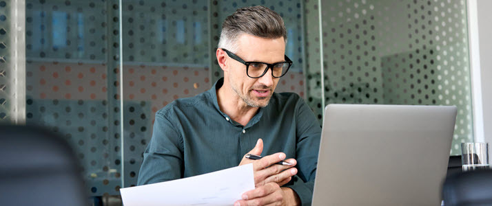 mature businessman at desk looking at computer with paper mature businessman at desk looking at computer with paper