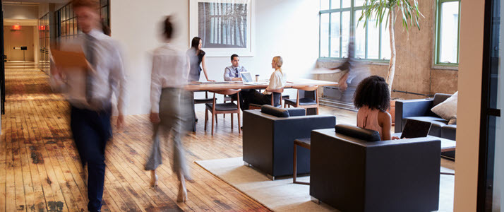 Employees walk through an office lobby where people sit in armchairs and at a conference table