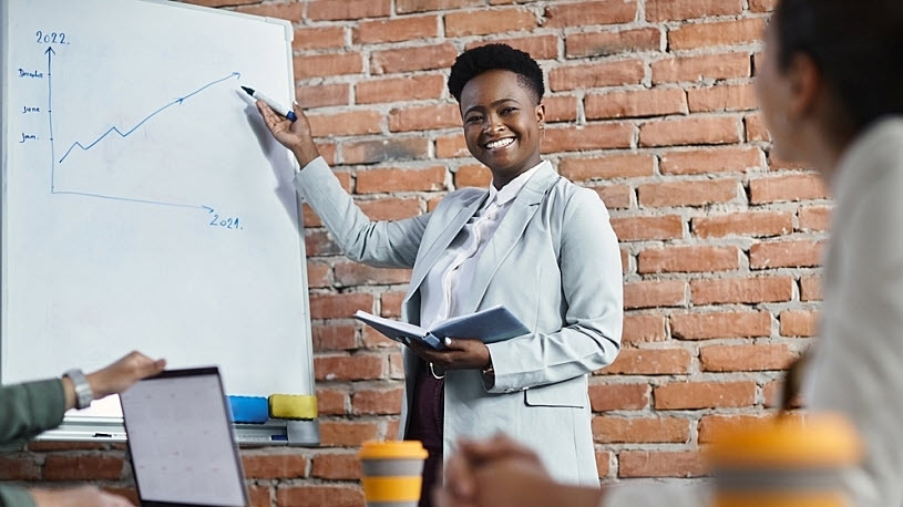 A Black businesswoman holds a notebook in one hand and points to whiteboard while speaking to group