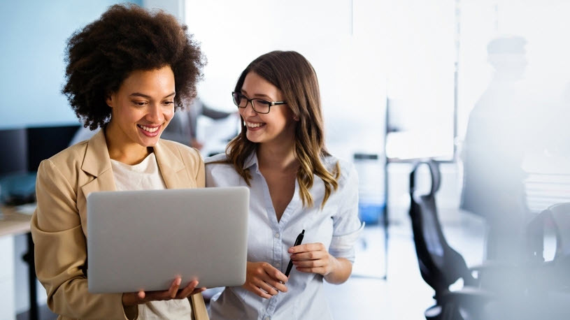 Pay transparency compliance Two women colleagues chat in an office while one looks at a laptop