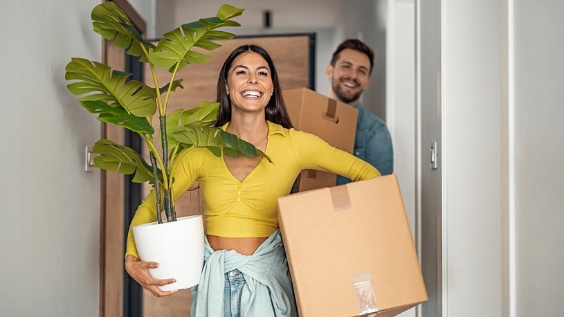 Compliance Trends Smiling woman and man carry moving boxes and plants out of apartment