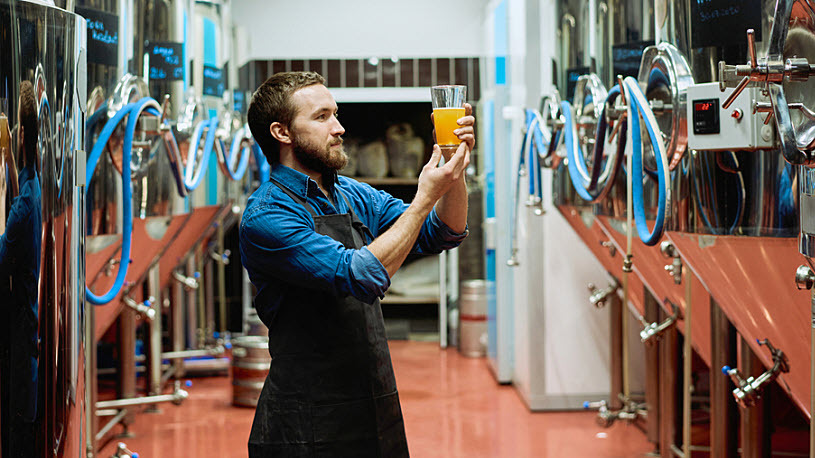 Young brewery worker examines beer sample among storage tanks Young brewery worker examines beer sample among storage tanks observing workplace safety rules