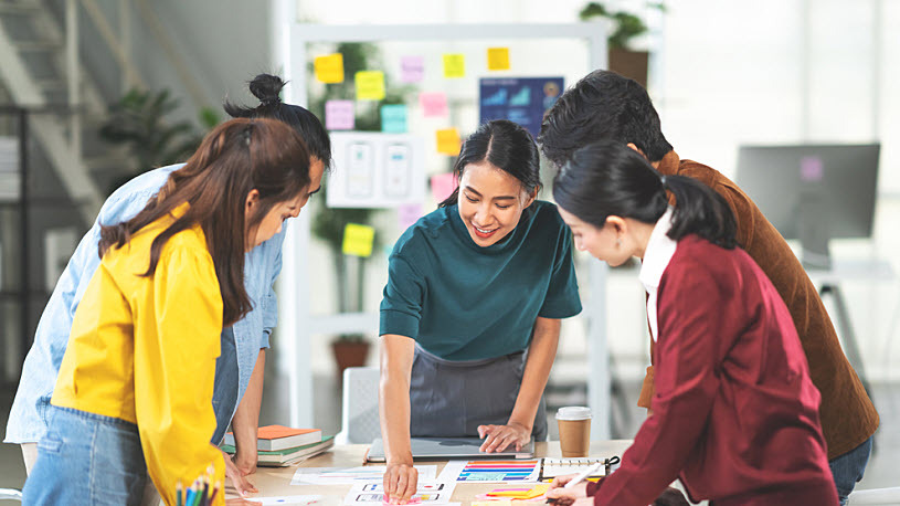 Employee Experience Management Five workers stand around a table in an office looking at papers together.