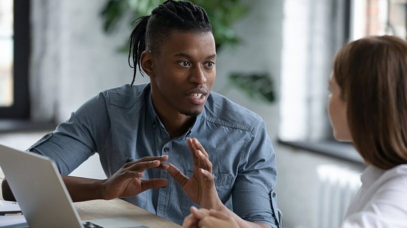AI for HR: How It Benefits Your Business (And How to Get Buy-In) A young man sitting at desk with laptop gestures and talks to coworker