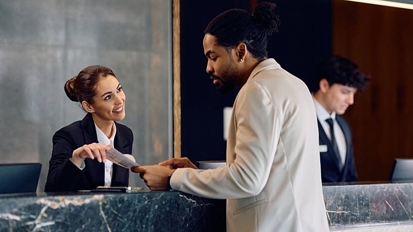 HR Technology in the Hospitality Industry in 2025 Hotel receptionist in uniform at a desk in the lobby assisting guest