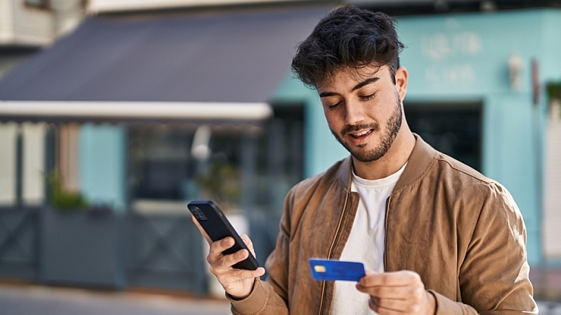 Pros and Cons of Payroll Card Programs: Are Paycards Better? Smiling man is using smartphone and payroll card for paying in e-shop