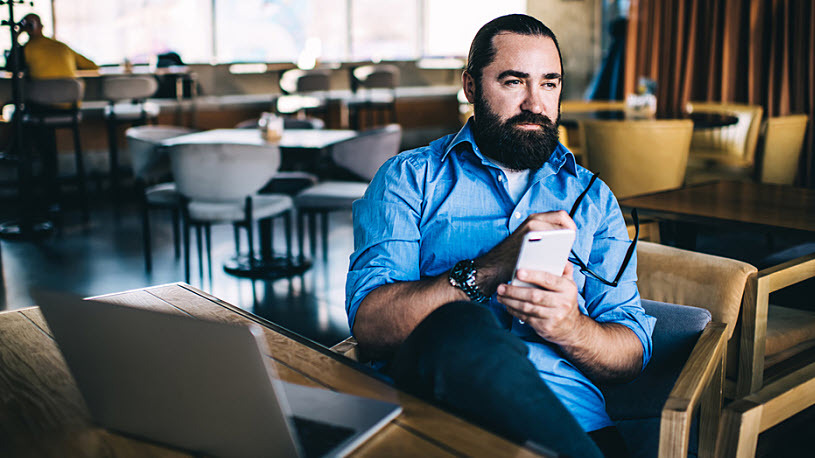 A serious bearded man in a cafe thinking with phone and laptop A serious bearded man in a cafe sits thinking with a phone and laptop