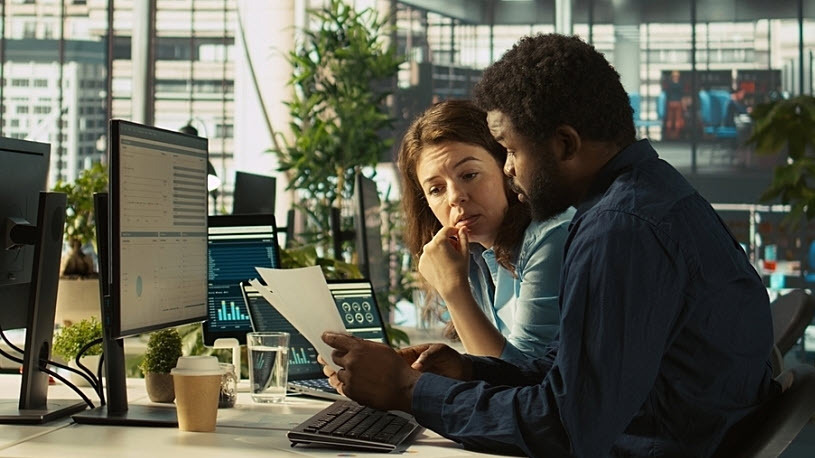Manager sits at desk reviewing reports with team member