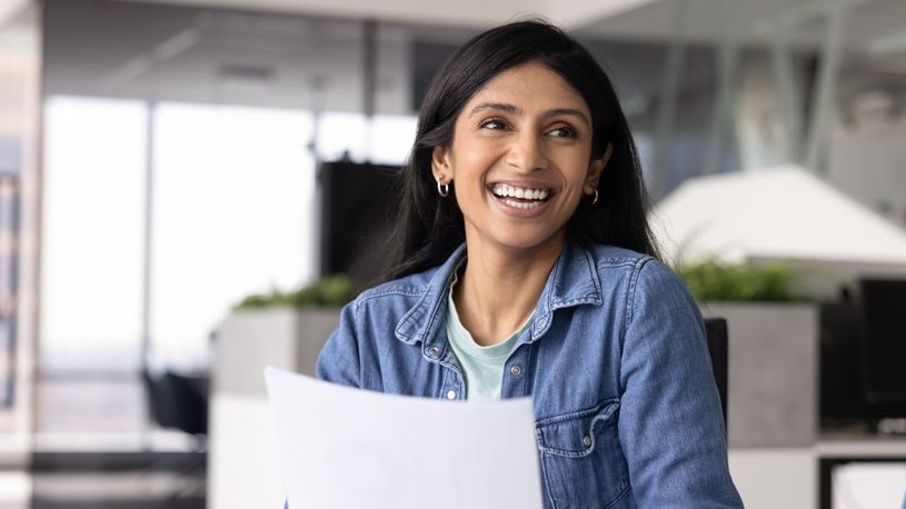 A young professional smiles confidently in an office setting while holding a stack of papers, representing the early career experience of new graduates entering the workforce.