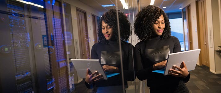 A millennial woman leans against a glass partition and holds a laptop
