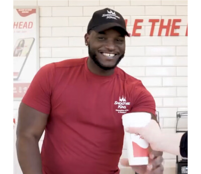 Smoothie King employee smiling while handing a drink to a customer behind the counter.