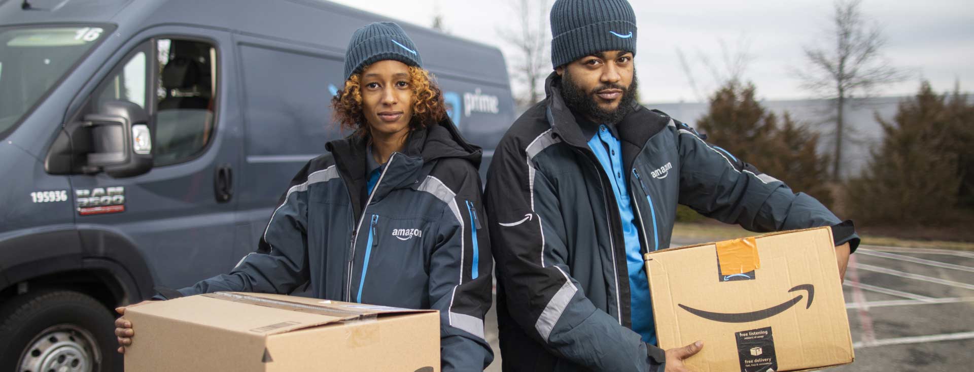 Two Amazon delivery people each holding Amazon boxes in front of delivery van.